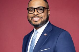 Black man in glasses and a blue suit smiling in front of a red background